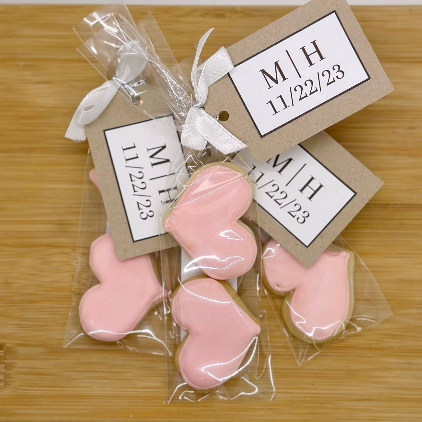 Pink heart-shaped cookies in clear packaging with white bows and personalized tags that say "M&H 11/22/23" sit on a wooden tray.