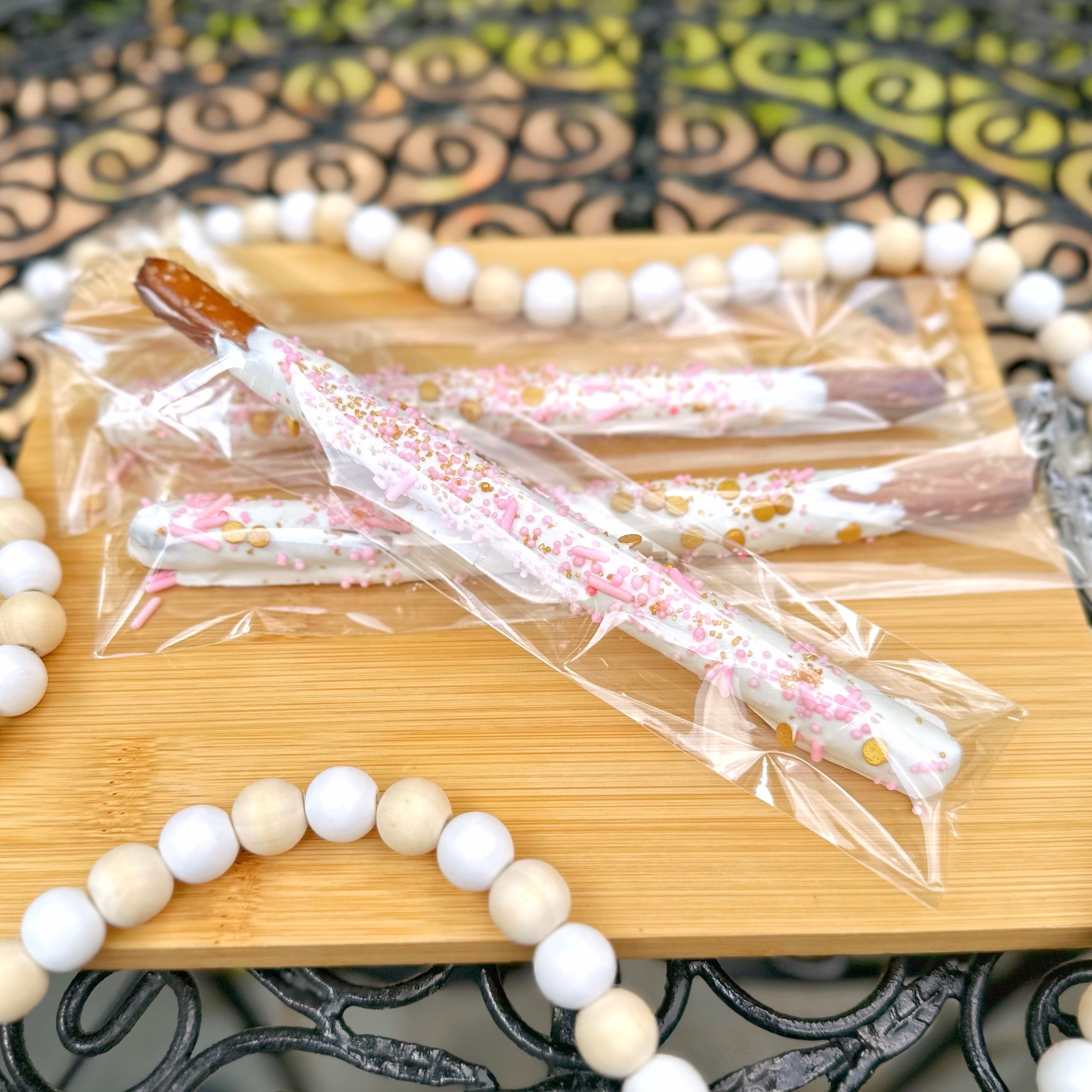 Three dipped pretzel rods decorated with white candy melts and pink and gold sprinkles sits on a wooden tray with a wooden beaded string around it.