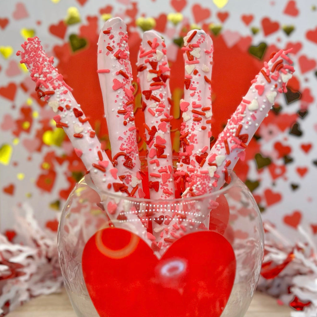 Dipped pretzel rods with light pink candy melts and red and white heart sprinkles in a glass vase against a heart-themed background.