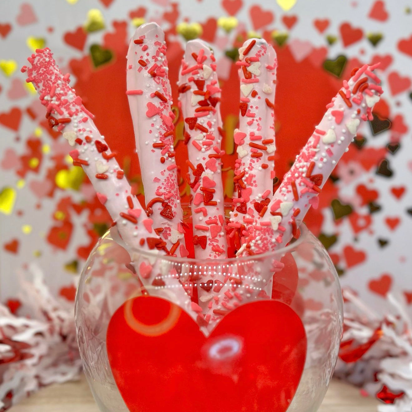 Dipped pretzel rods with light pink candy melts and red and white heart sprinkles in a glass vase against a heart-themed background.