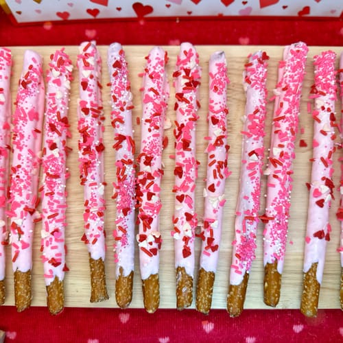 Dipped pretzel rods with light pink candy melts and pink and red heart sprinkles on a wooden board with a red background.