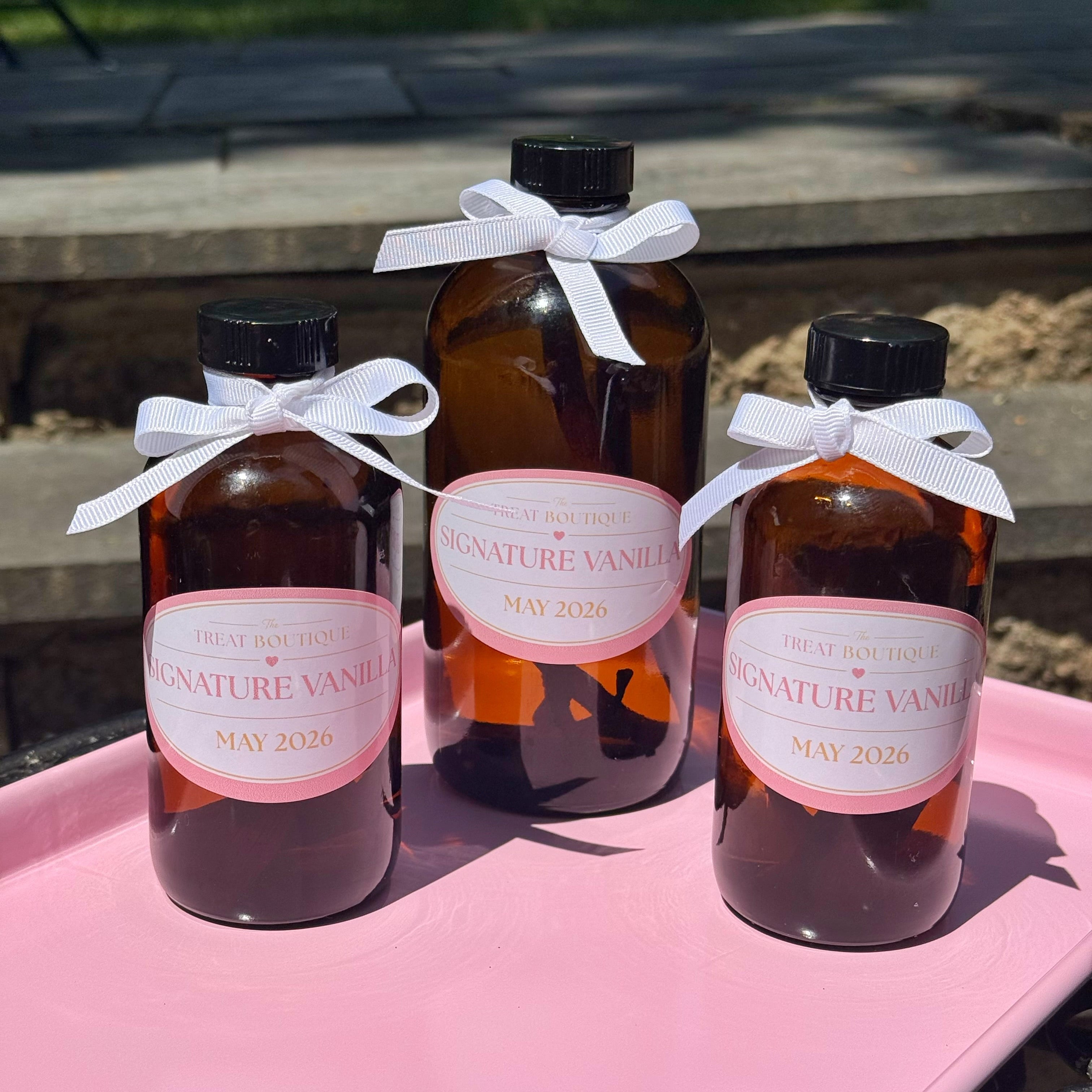 Three amber glass bottles with white labels and ribbons on a pink surface outdoors.
