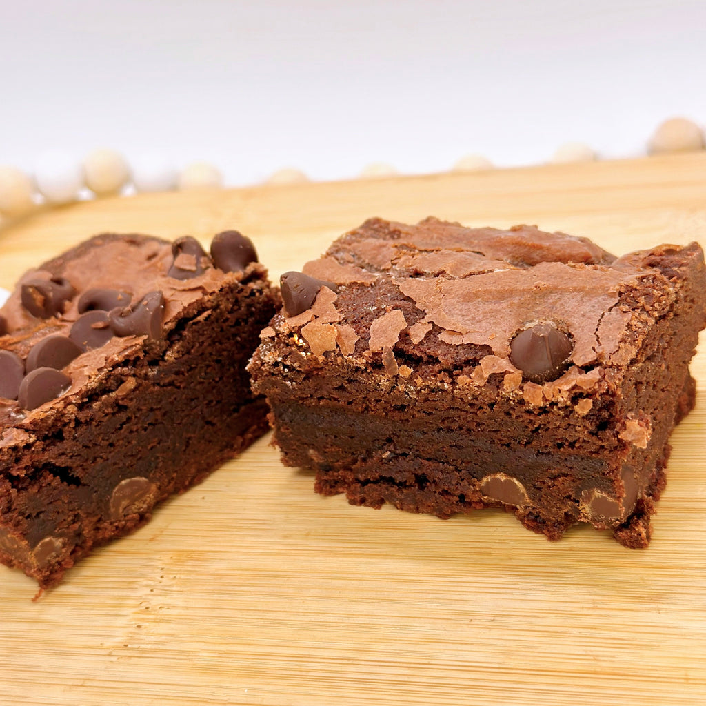 A close up of a chocolate brownie with chocolate chips cut in half on a wooden board, surrounded by a white beaded string.