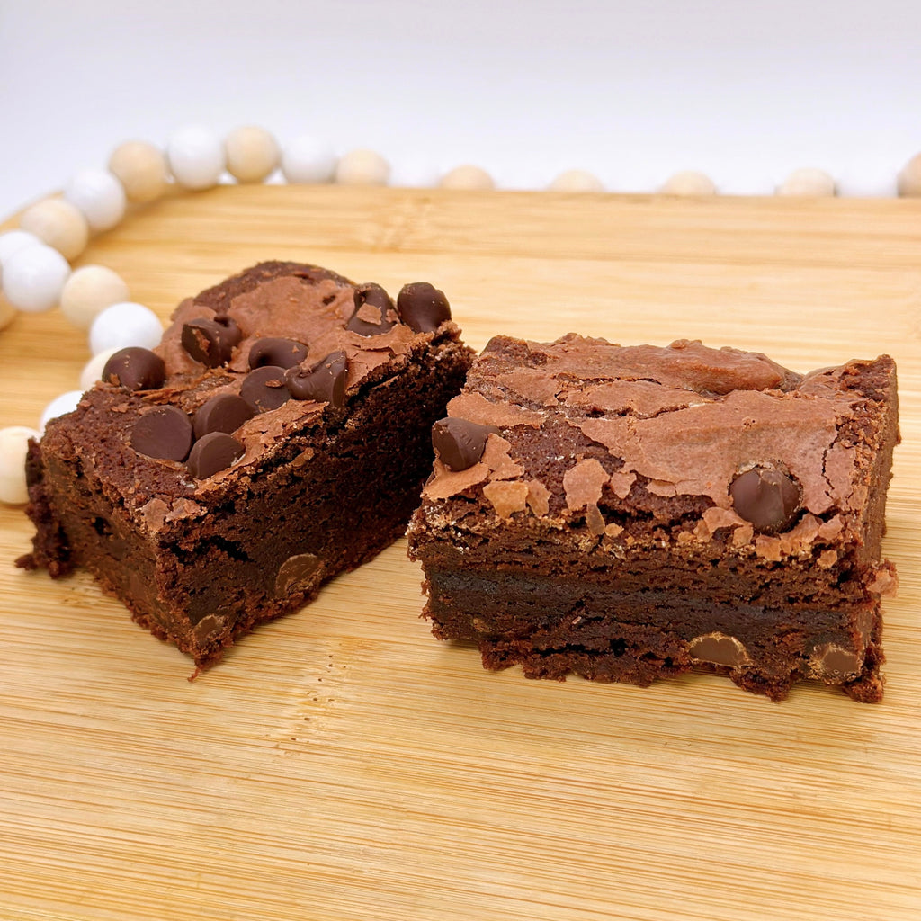 A chocolate brownie with chocolate chips cut in half on a wooden board, surrounded by a white beaded string.