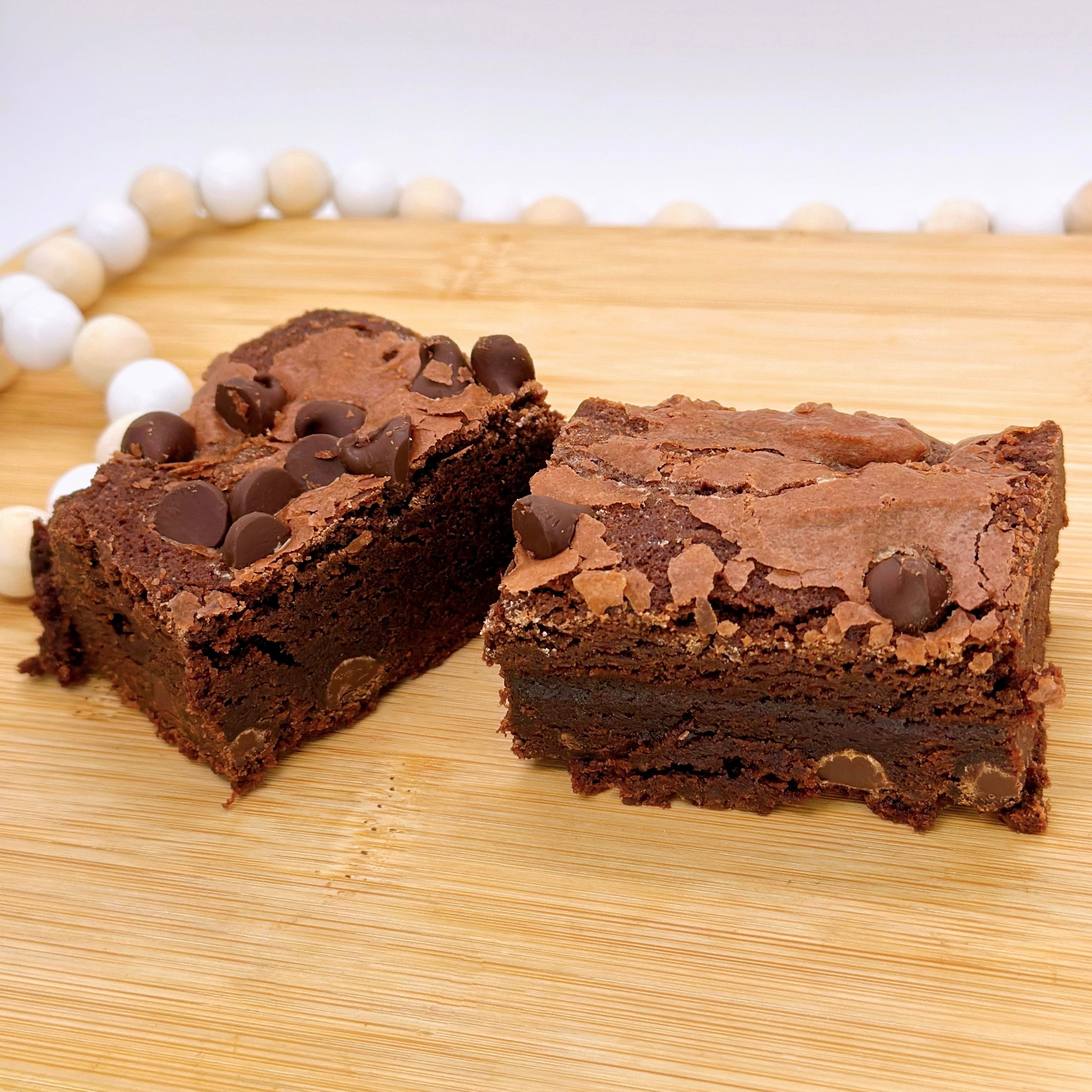 A chocolate brownie with chocolate chips cut in half on a wooden board, surrounded by a white beaded string.
