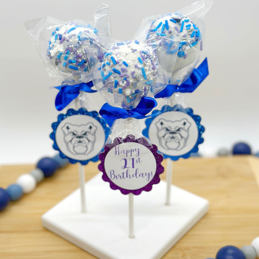Three cake pops decorated in blue and purple with blue bows and Butler bulldog themed tags and one purple sparkly tag that says "Happy 21st Birthday" on it sit on a wooden tray with a blue and white beaded string around it.