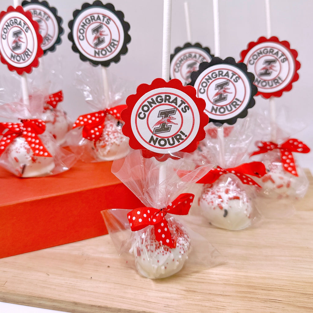 black and red themed cake pops with red polka dot bows and "Congrats Nour!" tags in black and red sit on a wooden tray.