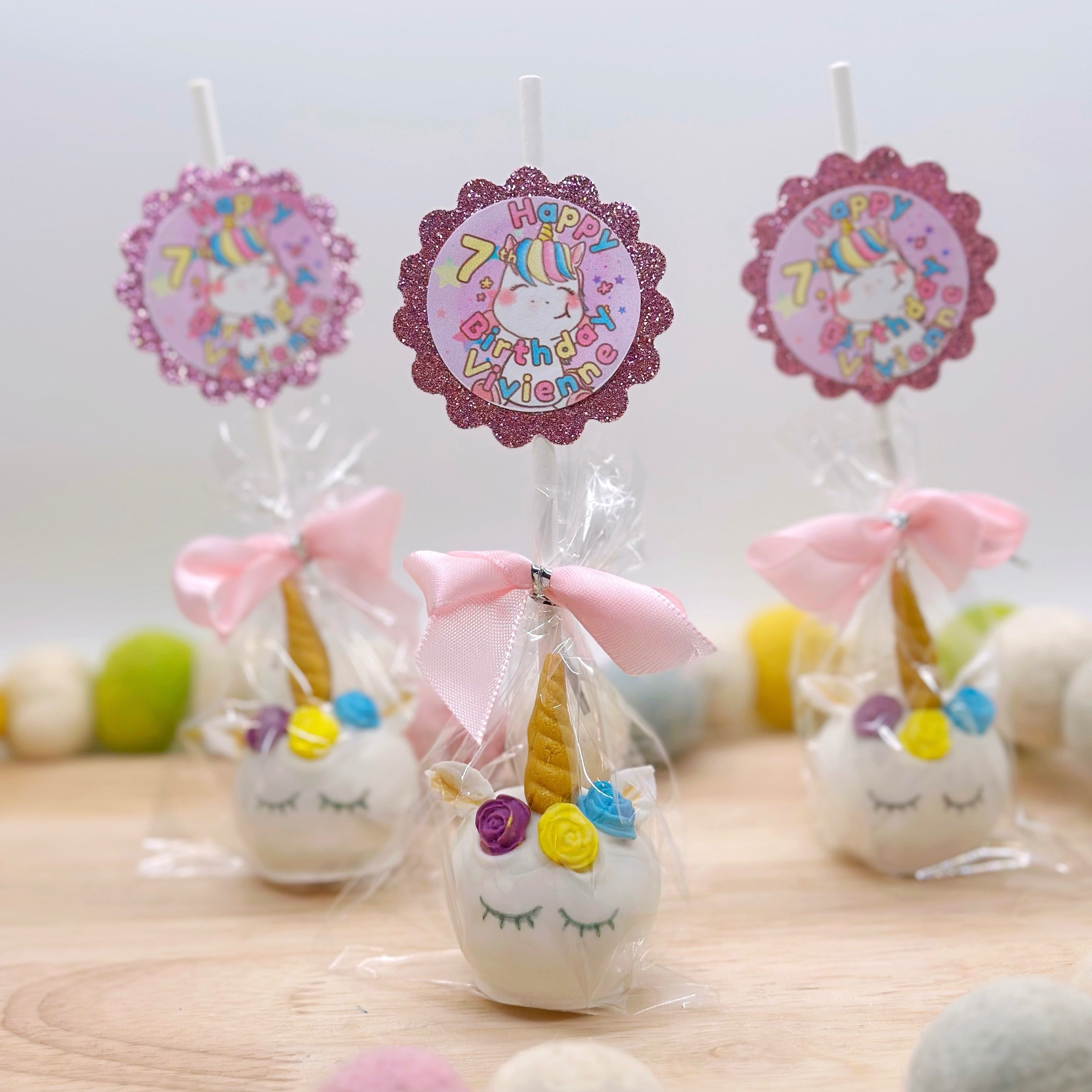 Birthday cake pops in the shape of unicorns with pink sparkly tags that say "Happy 7th Birthday Vivienne" in rainbow colors with a unicorn in the background sit on a wooden tray.
