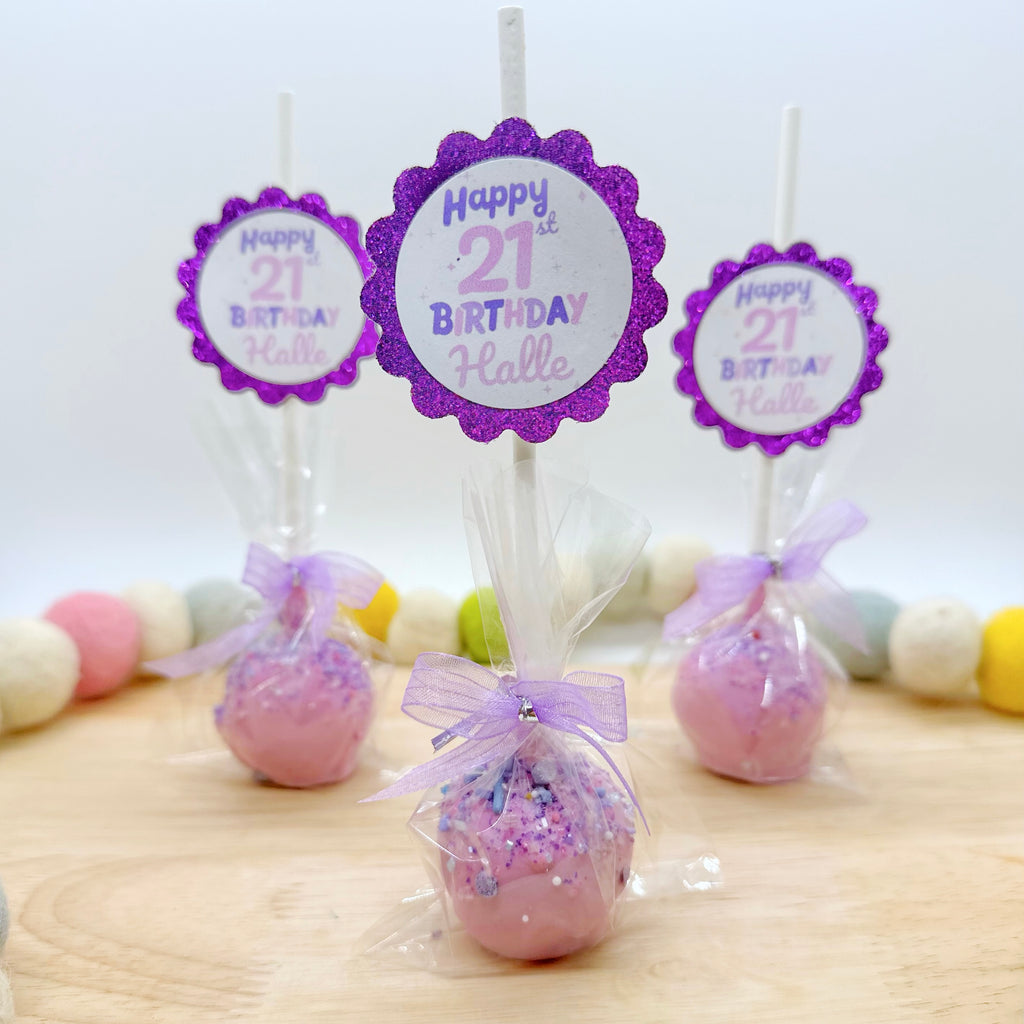 Three purple cake pops with light purple tags and purple sparkly tags that say " Happy 21st birthday Halle" on them in purple sit on a wooden tray with a multicolor felt beaded string around it.