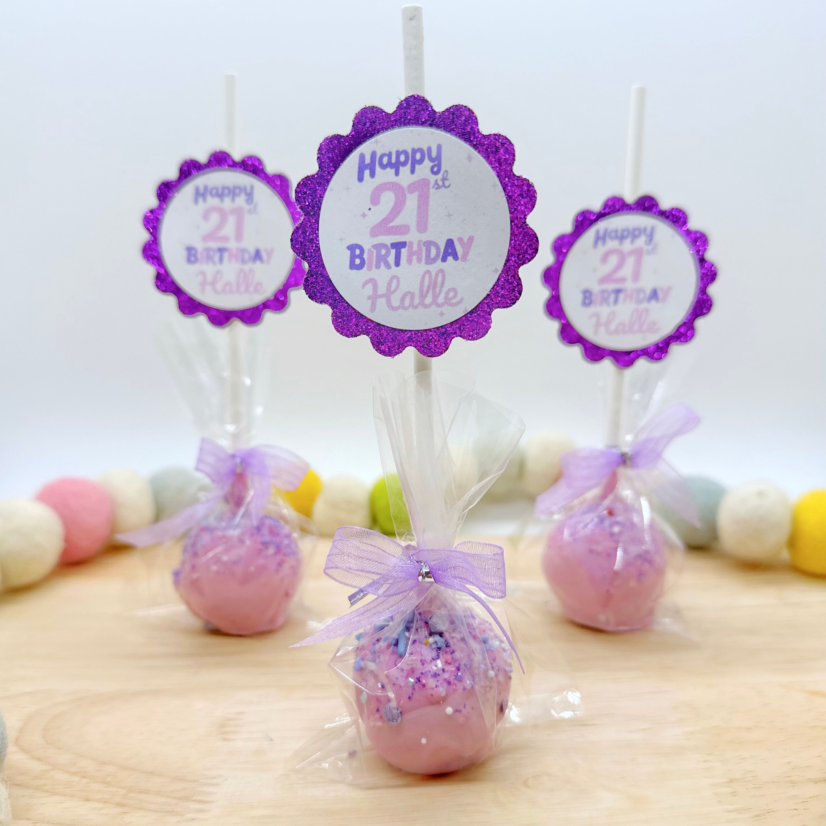 Three purple cake pops with light purple tags and purple sparkly tags that say " Happy 21st birthday Halle" on them in purple sit on a wooden tray with a multicolor felt beaded string around it.