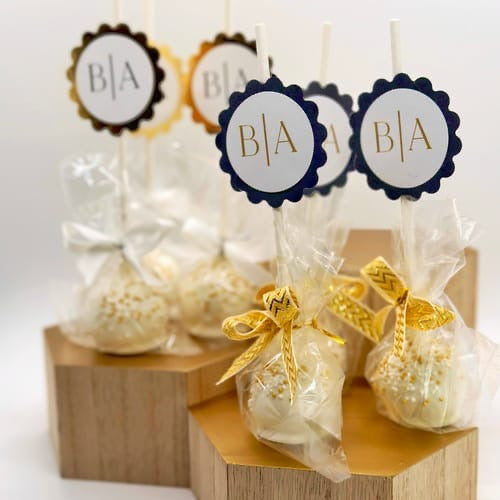 gold and white cake pops with gold tags that say "B&A" with gold ribbons sit on stacked hexagonal blocks that are wooden with gold tops.
