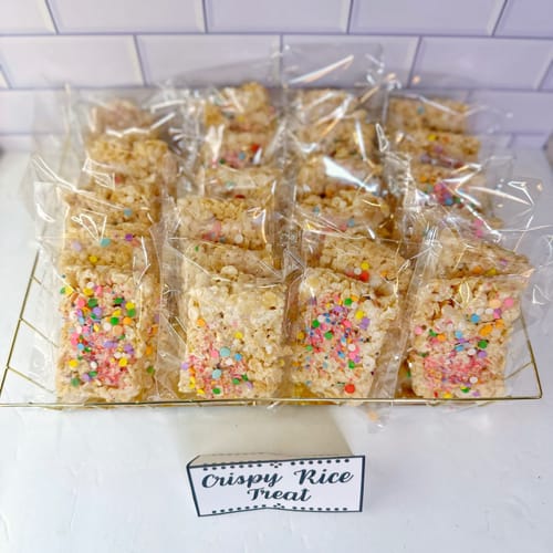 Crispy rice treats with rainbow sprinkles in clear packaging on a white surface with a tiled wall background.