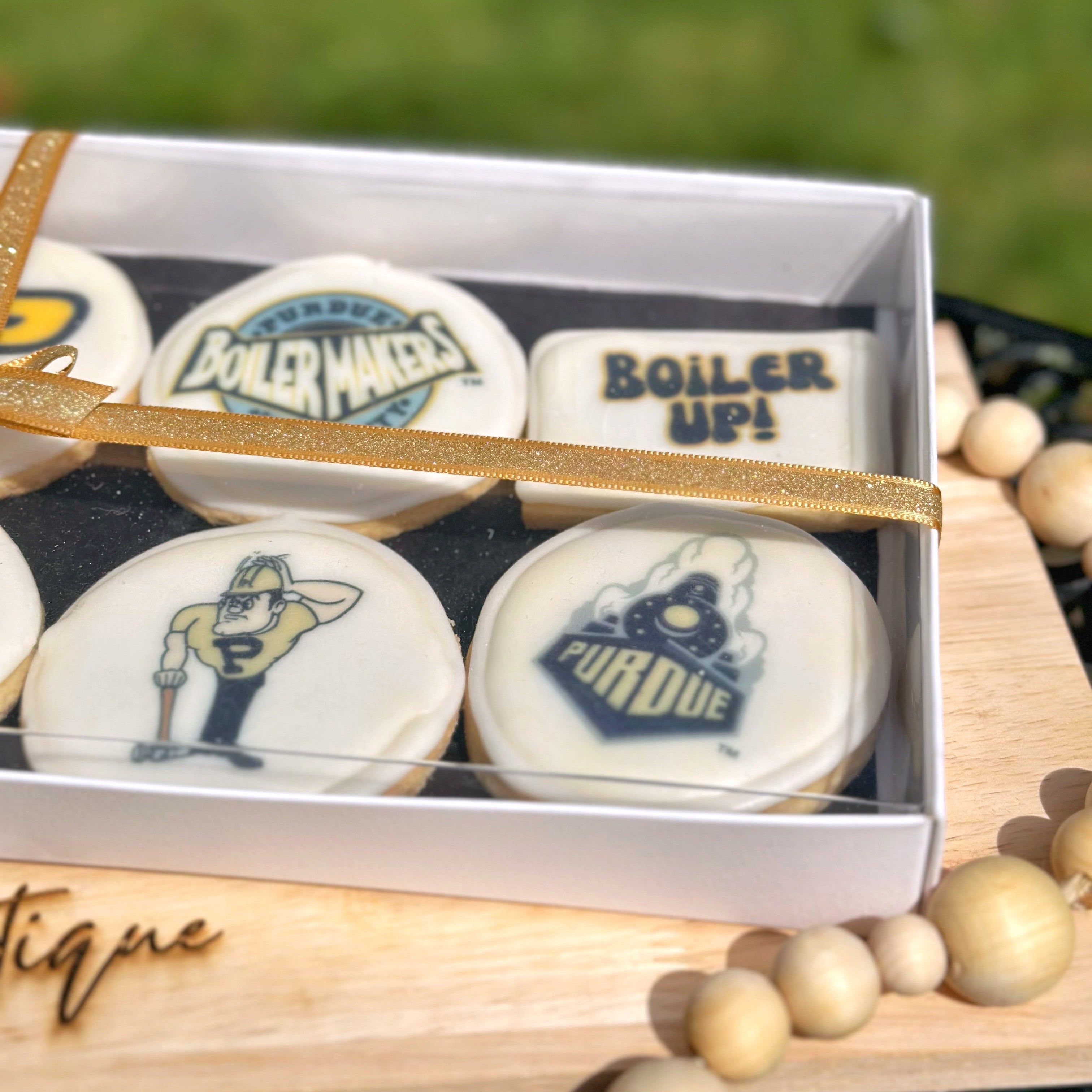 A different angle of a Purdue themed cookie set with a gold ribbon on a wooden stand, branded 'The Treat Boutique', with a blurred grass background.