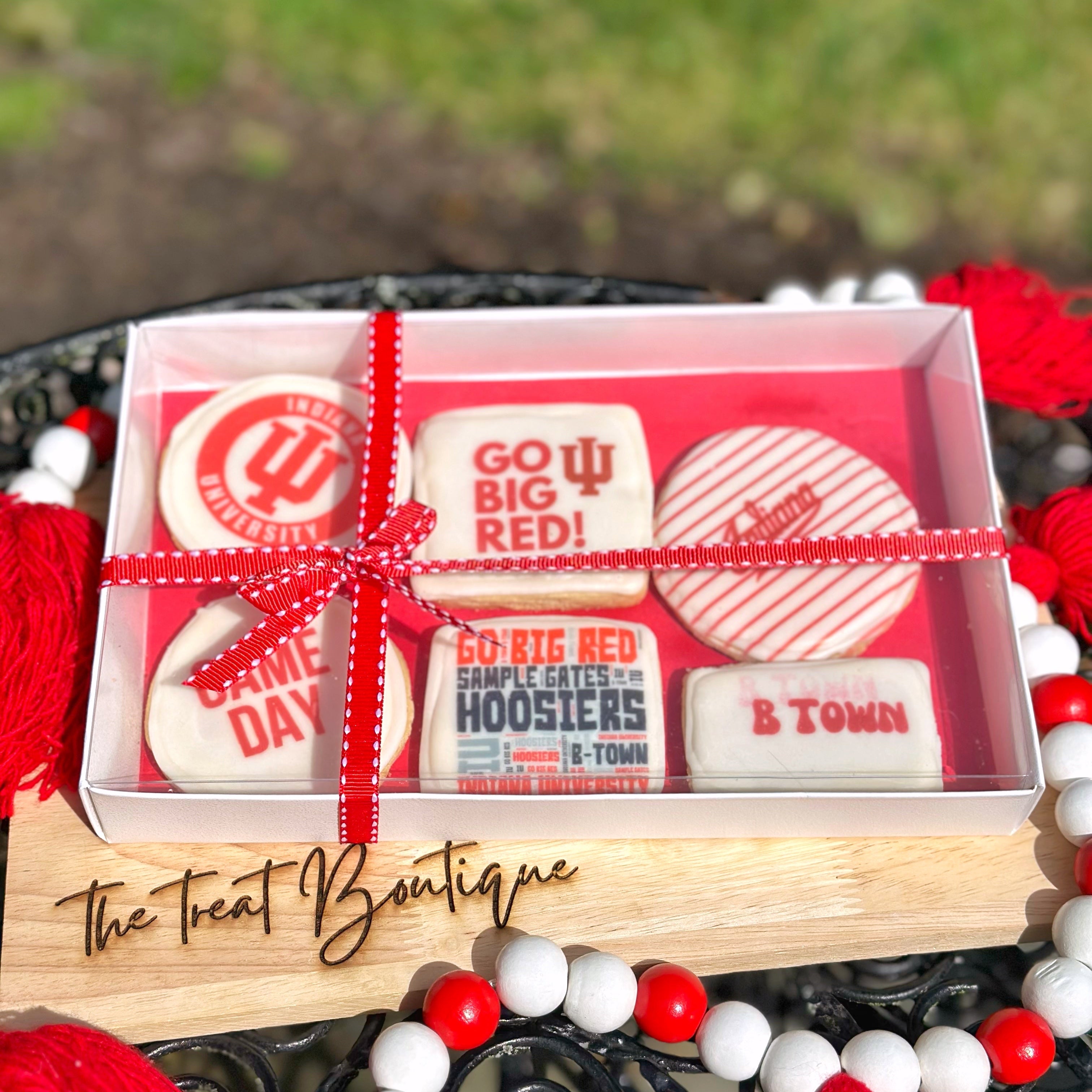 An IU themed cookie set with a red ribbon on a wooden stand, branded 'The Treat Boutique', with a blurred grass background.