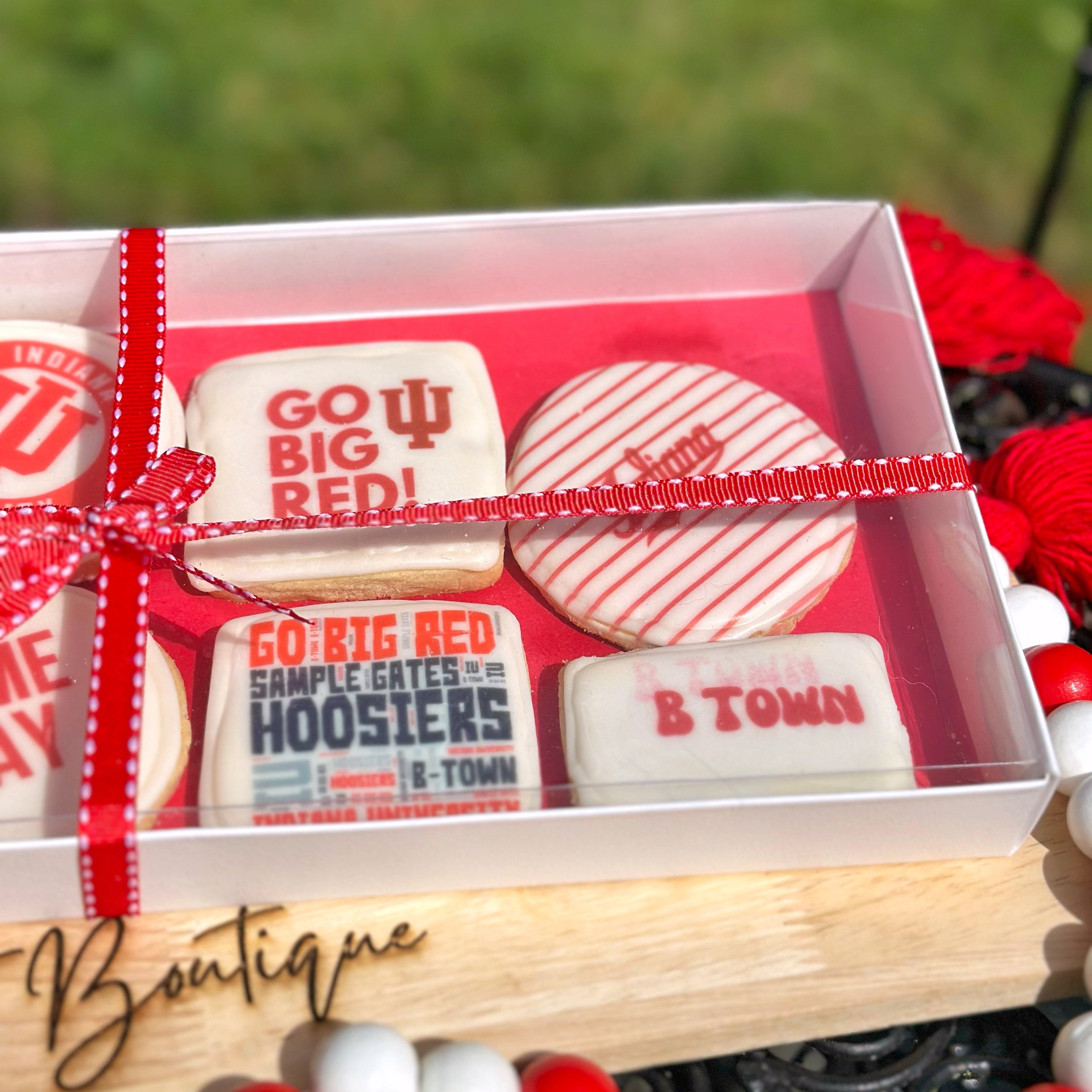Close up of an IU themed cookie set with a red ribbon on a wooden stand, branded 'The Treat Boutique', with a blurred grass background.