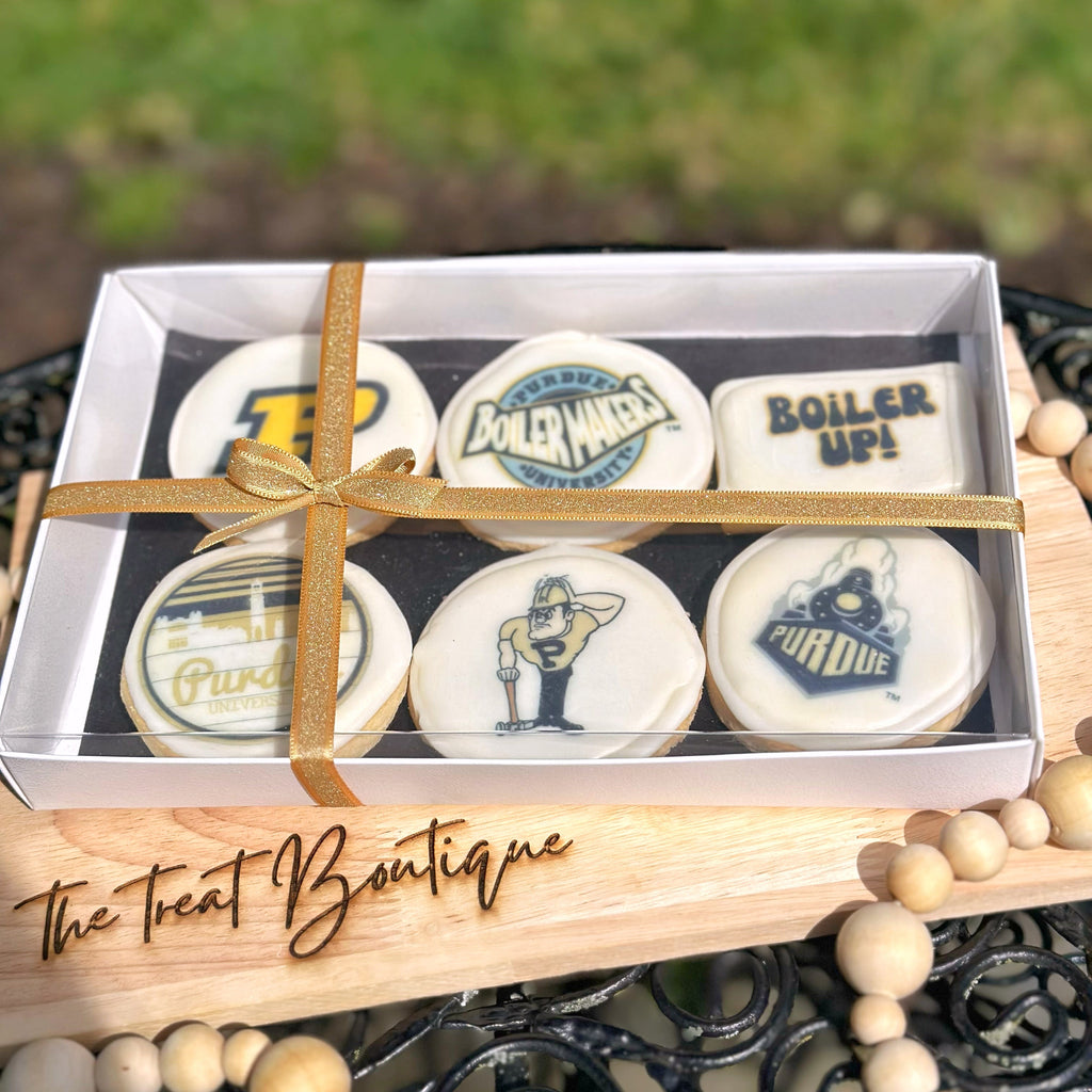 An Purdue themed cookie set with a gold ribbon on a wooden stand, branded 'The Treat Boutique', with a blurred grass background.