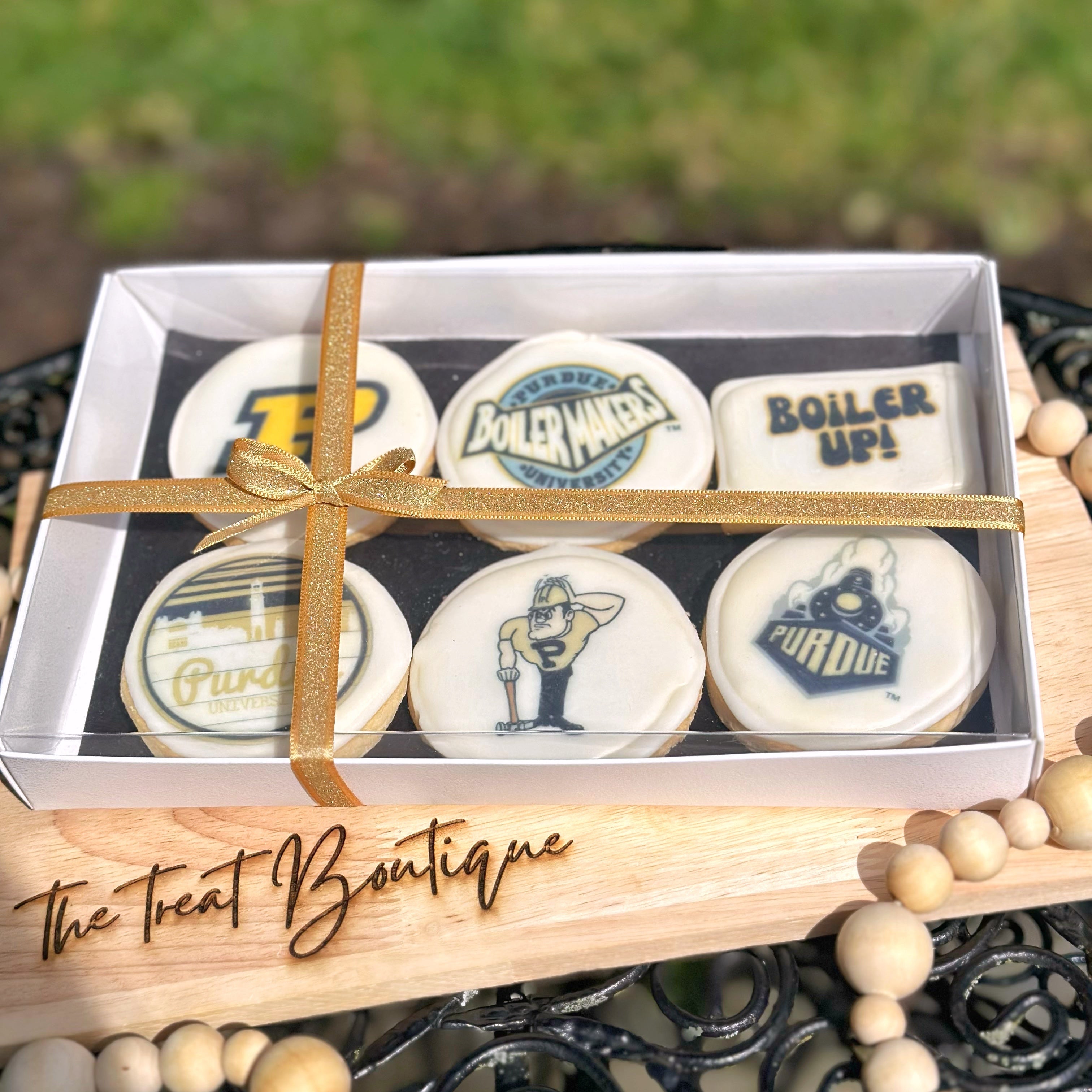 An Purdue themed cookie set with a gold ribbon on a wooden stand, branded 'The Treat Boutique', with a blurred grass background.