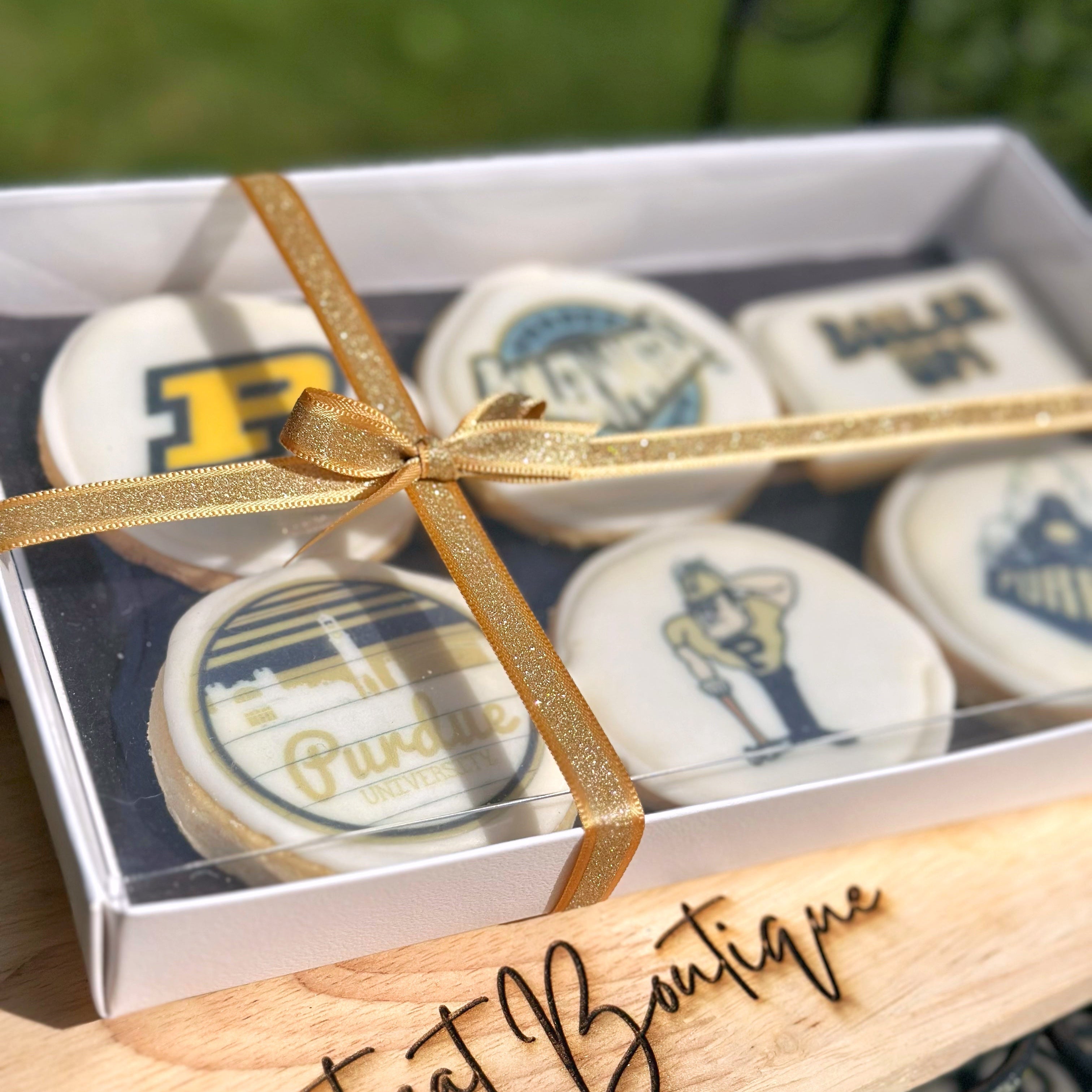 A close up of a Purdue themed cookie set with a gold ribbon on a wooden stand, branded 'The Treat Boutique', with a blurred grass background.