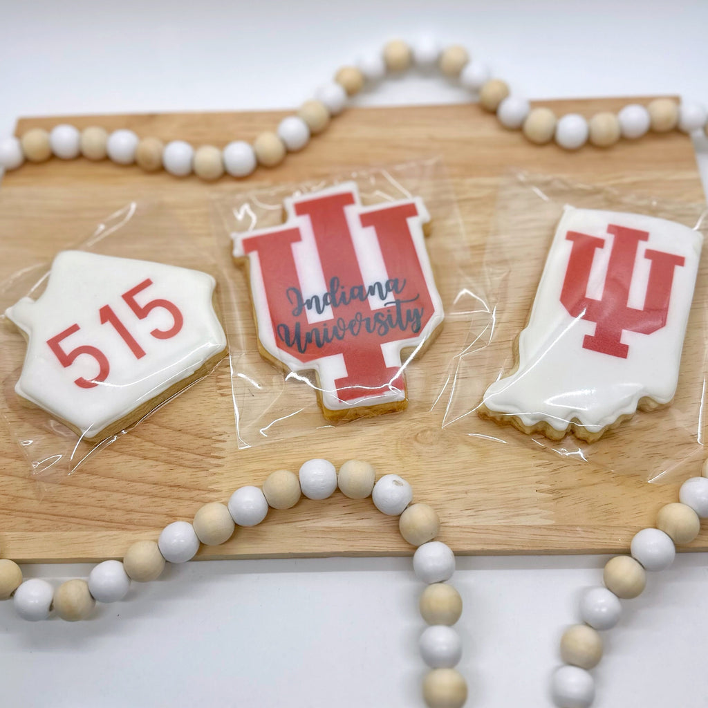 Three cookies sit on a wooden tray with a wooden beaded string around it. One cookie is shaped like a house with 515 printed on it in red. One cookie is the shape of the IU logo with the logo printed in red and "Indianapolis University" written atop it in black cursive. One cookie is the shape of the state of Indiana with IU logo printed on it in red.