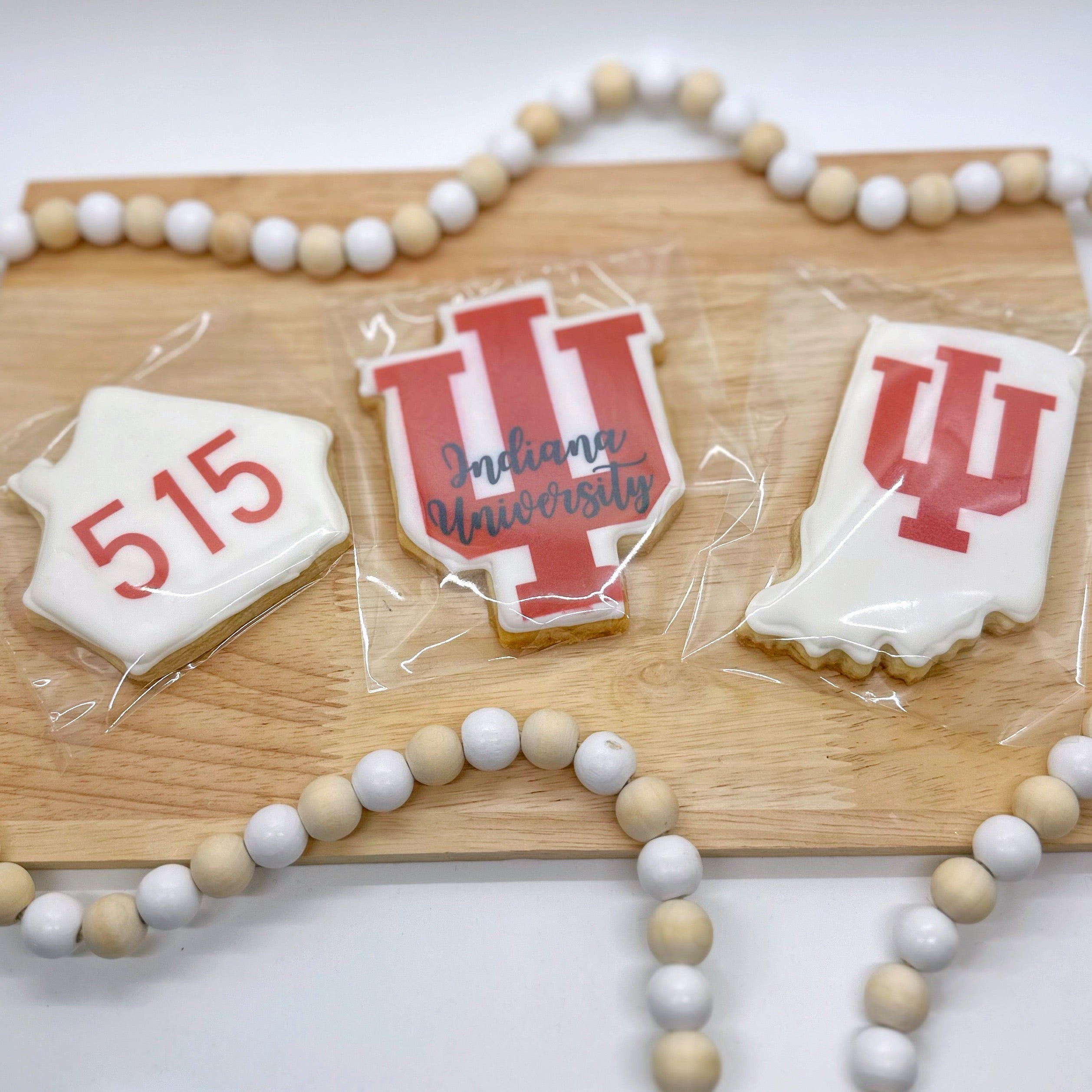 Three cookies sit on a wooden tray with a wooden beaded string around it. One cookie is shaped like a house with 515 printed on it in red. One cookie is the shape of the IU logo with the logo printed in red and "Indianapolis University" written atop it in black cursive. One cookie is the shape of the state of Indiana with IU logo printed on it in red.