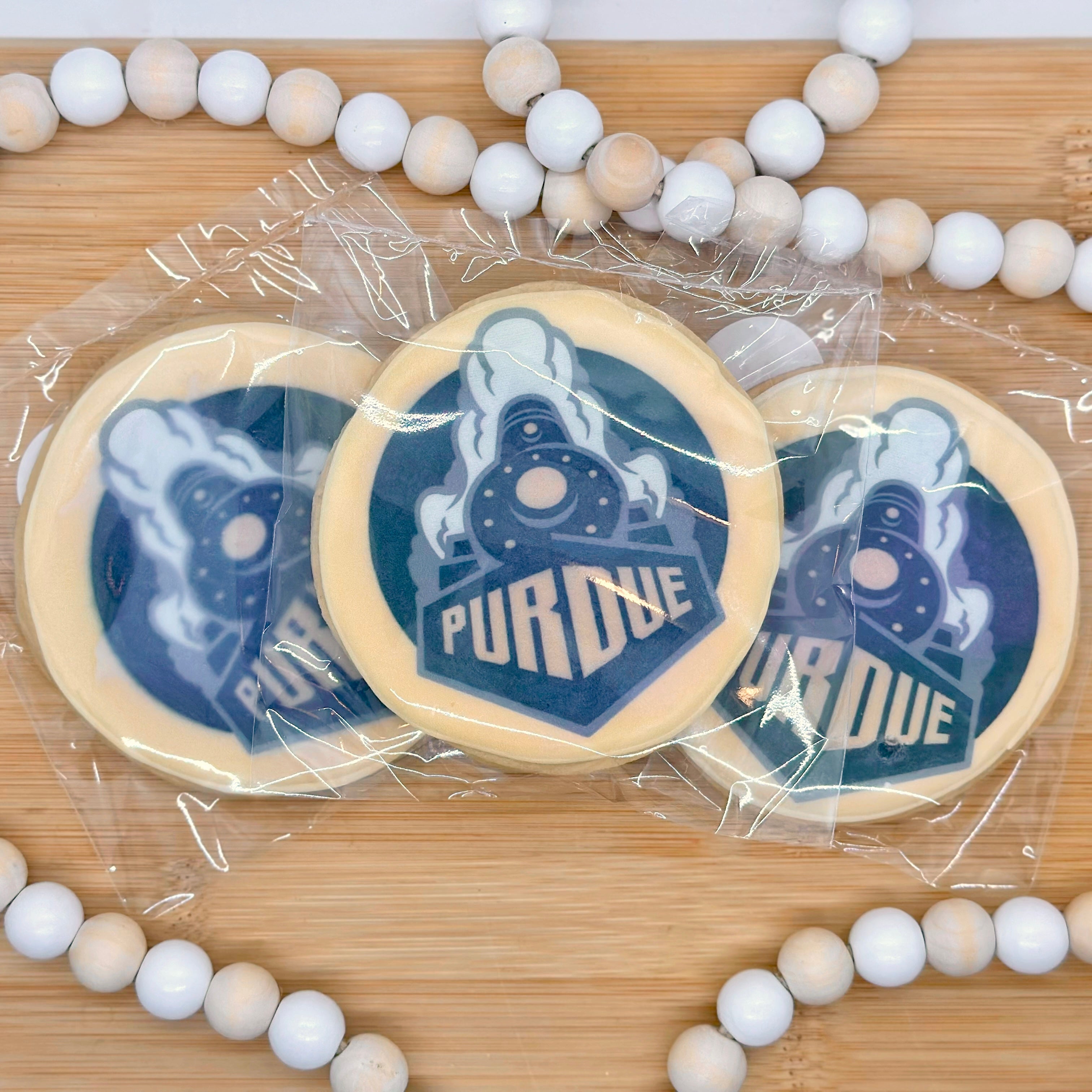Three circle cookies with the Purdue logo printed on it in black and gold sit on a wooden tray with a wooden beaded string around it.