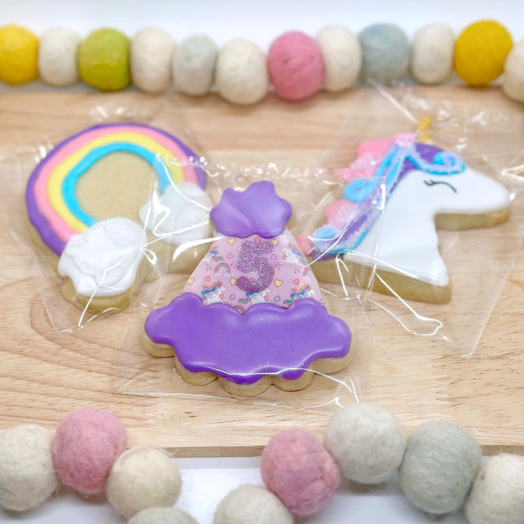 Decorative cookies shaped like a rainbow, a party hat with a sparkly number 5, and rainbow unicorn on a wooden tray with a colorful felt bead string around it.