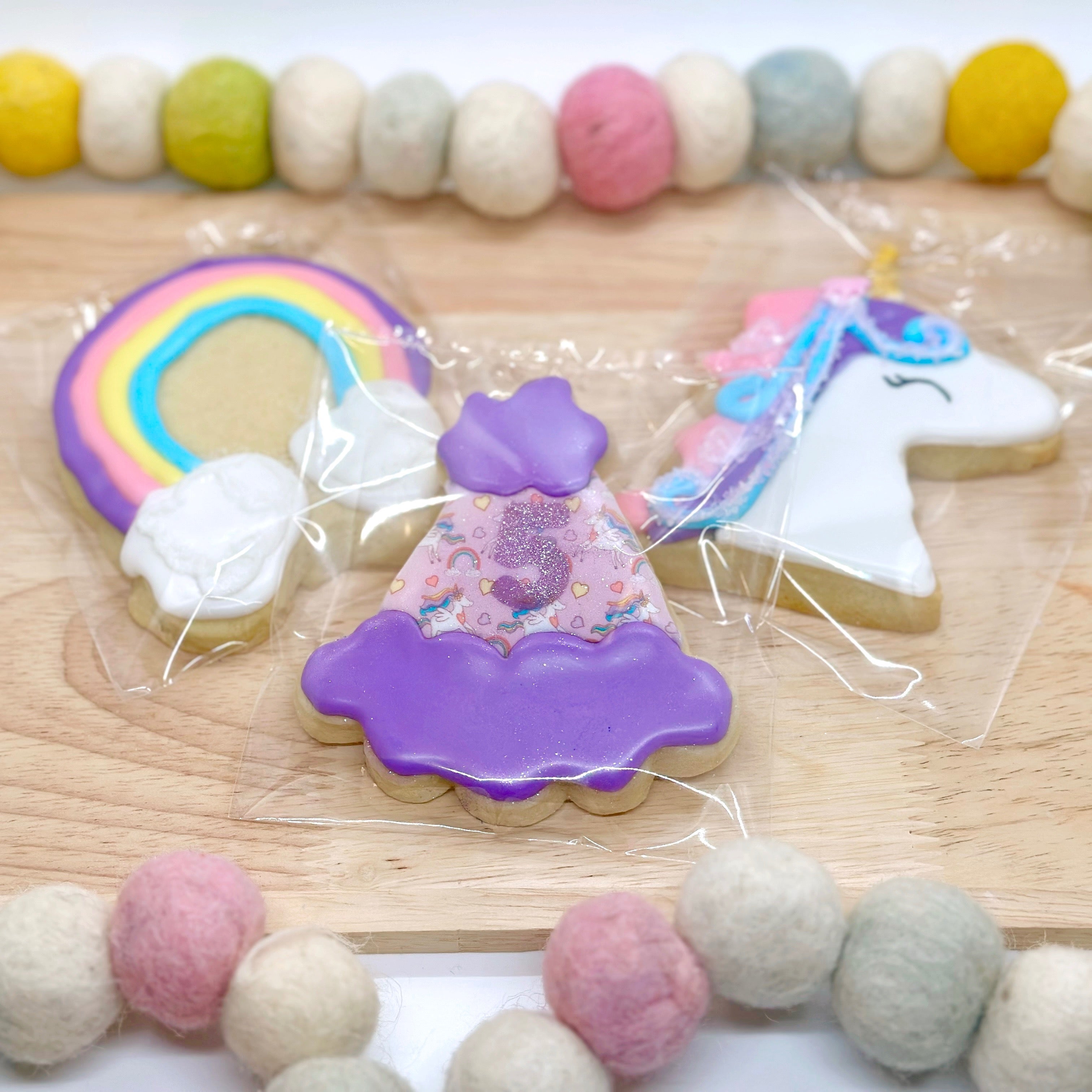 Decorative cookies shaped like a rainbow, a party hat with a sparkly number 5, and rainbow unicorn on a wooden tray with a colorful felt bead string around it.