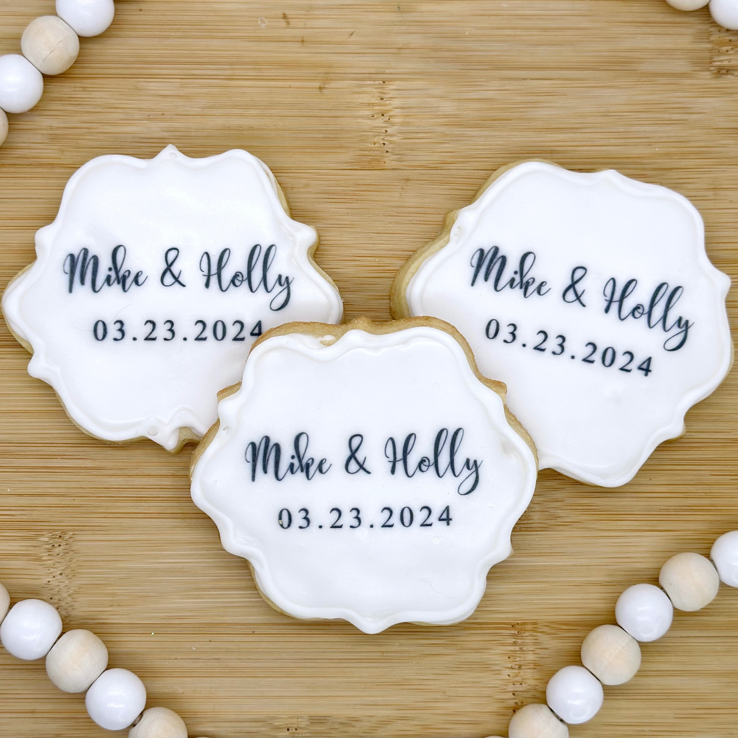 Three scalloped cookies with "Mike & Holly 03/23/24" printed on them in black sit on a wooden tray with a beaded wooden string around it.