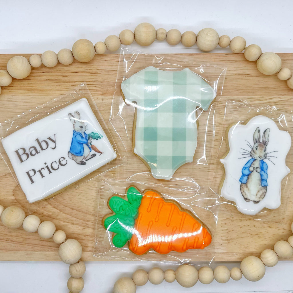 4 cookies sit on a wooden tray with a wooden beaded string around them. One cookie is a rectangle shape with the photo of a bunny on it and the text "baby price" in brown. One cookie is shaped like a onesie printed with a pale green plaid. One cookie is a plump orange carrot with a green stem. The Last cookie is a long scalloped rectangle with a different picture of a bunny on it.
